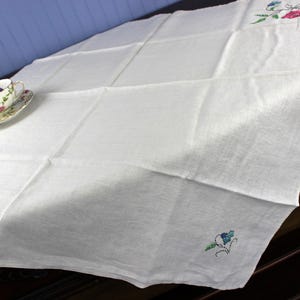 May include: White linen tablecloth with embroidered floral accents in pink, blue, and green. A floral teacup and saucer sit on the tablecloth. The tablecloth is folded and draped over a dark wooden surface.