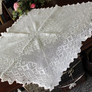 May include: White crocheted tablecloth with a delicate floral pattern. The tablecloth is laid out on a wooden table.