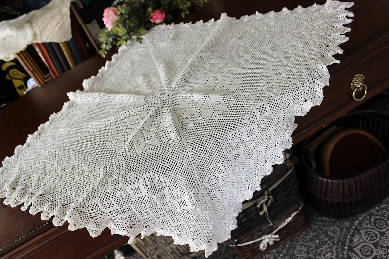 May include: White crocheted tablecloth with a delicate floral pattern. The tablecloth is laid out on a wooden table.