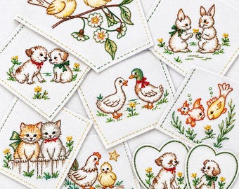 May include: A collection of white fabric patches featuring embroidered illustrations of pairs of animals including birds, rabbits, puppies, kittens, ducks, fish, and a hen with a chick. Some patches are square with stitched borders, and two are heart-shaped.