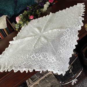 May include: A white crocheted tablecloth with a delicate lace pattern. The tablecloth is laid out on a dark wooden table.