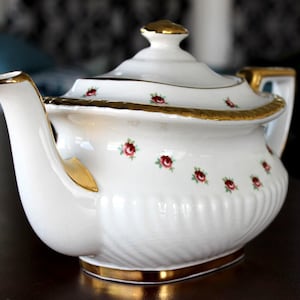 May include: White porcelain teapot with gold trim and accents. The teapot features a lid and handle with gold detailing. Small red rose floral pattern adorns the body of the teapot. The teapot is a classic design.