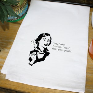 May include: A white tea towel with a black and white illustration of a woman drinking coffee. The text on the towel reads "Yes, I sew. And no, I won't hem your pants."