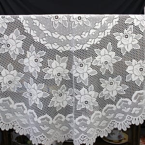 May include: White lace tablecloth with a floral pattern. The tablecloth is rectangular and features a delicate, openwork design with repeating flower motifs. The edges are scalloped, adding a decorative touch to the table covering.