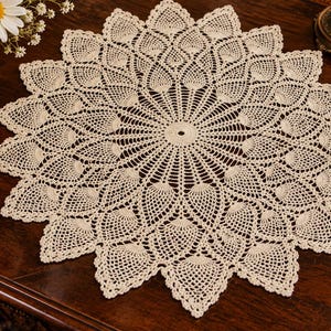 May include: A round, off-white crochet doily featuring a complex, radiating pattern of interconnected leaf-like motifs. The design starts from a solid circular center and expands outward into a series of pointed, scalloped edges along the perimeter.