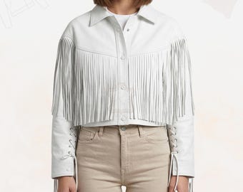 May include: A white cropped jacket with fringe detailing on the sleeves and chest. The jacket has a collared neckline, button-up front, and lace-up detailing on the sleeves. The model is wearing beige pants.