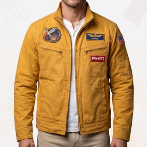 Può includere: Una giacca con cerniera color giallo senape dotata di colletto rialzato, due tasche sul petto con cerniere e varie toppe. Le toppe includono un design circolare a tema spaziale, un'etichetta blu con la scritta GRACE, un rettangolo rosso PHM e una piccola bandiera statunitense.