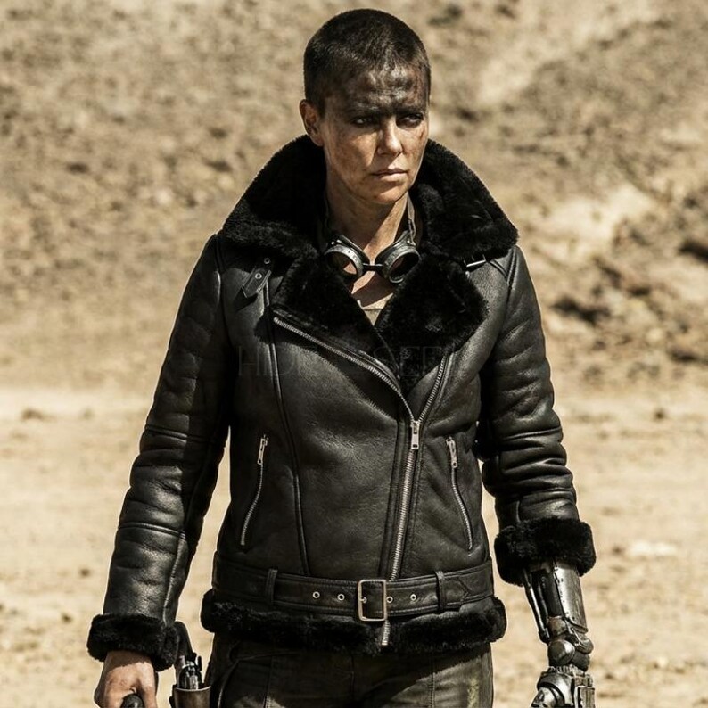 May include: A black leather jacket with a shearling collar and cuffs. The jacket features a belted waist, a zipper closure, and a pair of goggles around the neck. The image is set against a desert backdrop.