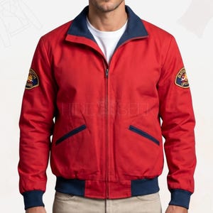 Peut inclure: Une veste zippée rouge avec un col, des poignets et un ourlet bleu marine. La veste comprend deux poches avant inclinées avec une bordure bleu marine et un écusson circulaire Baywatch Lifeguard sur chaque manche. Un t-shirt blanc est visible.