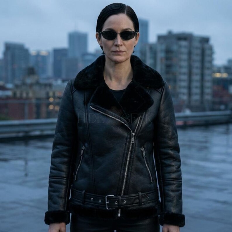 May include: Black leather jacket with a shearling collar and cuffs. The jacket features a diagonal zipper closure, a belted waist, and two front pockets. The jacket is worn over a black top and is paired with black pants. The background shows a cityscape.