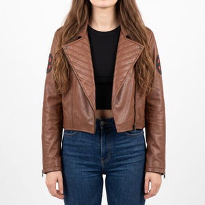 May include: A cropped brown leather jacket featuring wide lapels with diagonal quilted stitching, a black zipper closure, and circular black and red patches on both sleeves. The jacket is worn over a plain black top and blue denim jeans.