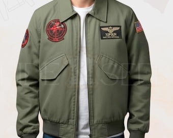 Könnte beinhalten: Olivgrüne Bomberjacke mit Reißverschluss, zwei Vordertaschen und gerippten Bündchen und Saum. Mit gestickten Aufnähern auf Brust und Ärmeln, darunter ein Top Gun-Logo und eine amerikanische Flagge. Die Jacke hat einen klassischen Kragen.
