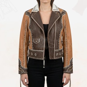 May include: Brown and tan leather jacket with decorative silver studs and embroidery. The jacket features a notched collar, a zip closure, and a fitted silhouette. The jacket has a western-inspired design with star and floral accents.