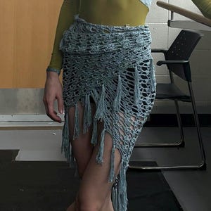 May include: A light blue crochet skirt with fringe detail. The skirt is made of a loosely woven material and has a textured appearance. The skirt is part of a dance or performance outfit.