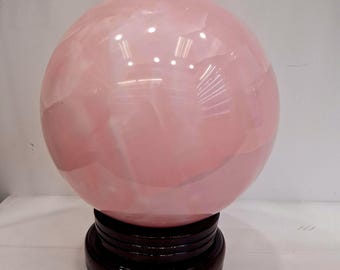 Könnte beinhalten: Eine große, polierte Rosenquarz-Kugel, die auf einem dunklen Holzständer ruht. Die Kugel hat eine zarte rosa Farbe mit subtilen inneren Adern, die das Licht reflektieren. Der Ständer ist dunkelbraun und mehrstufig.