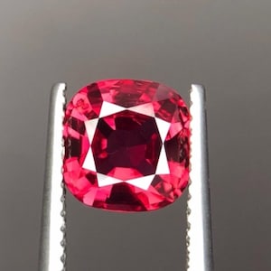 Pode incluir: Uma pedra preciosa facetada com corte almofada, segurada por pinças de metal. A gema é de cor vermelha vibrante com um centro quadrado e facetas triangulares brancas. O fundo é cinza sólido.