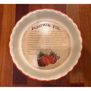 May include: A white pie dish with a red rim, featuring a recipe for pumpkin pie. The center of the dish has a tan background with the words "PUMPKIN PIE" and a recipe list. Two orange pumpkins are also depicted.