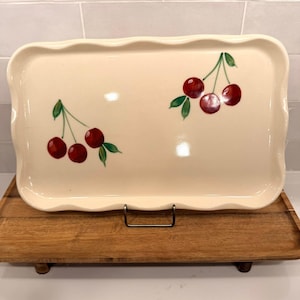 May include: Cream-colored rectangular serving platter with a scalloped edge. The platter is decorated with hand-painted red cherries and green leaves. The platter is displayed on a wooden tray.