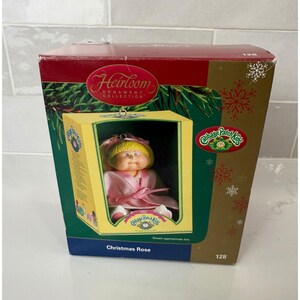 May include: A Cabbage Patch Kids Heirloom Ornament Collection box featuring a doll named Christmas Rose. The box is maroon and gold with snowflake graphics, displaying a yellow-framed window showing a doll with blonde hair in a pink robe.