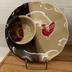 May include: A ceramic serving dish with a built-in dip bowl. The plate is tan and brown with a red rooster design. The dip bowl is brown and tan. The dish is on a metal stand.