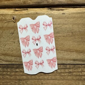 May include: A sheet of stickers featuring watercolor-style pink bows. The bows are in various sizes and arrangements, with a soft, delicate appearance. The stickers are on a white background with a scalloped edge.