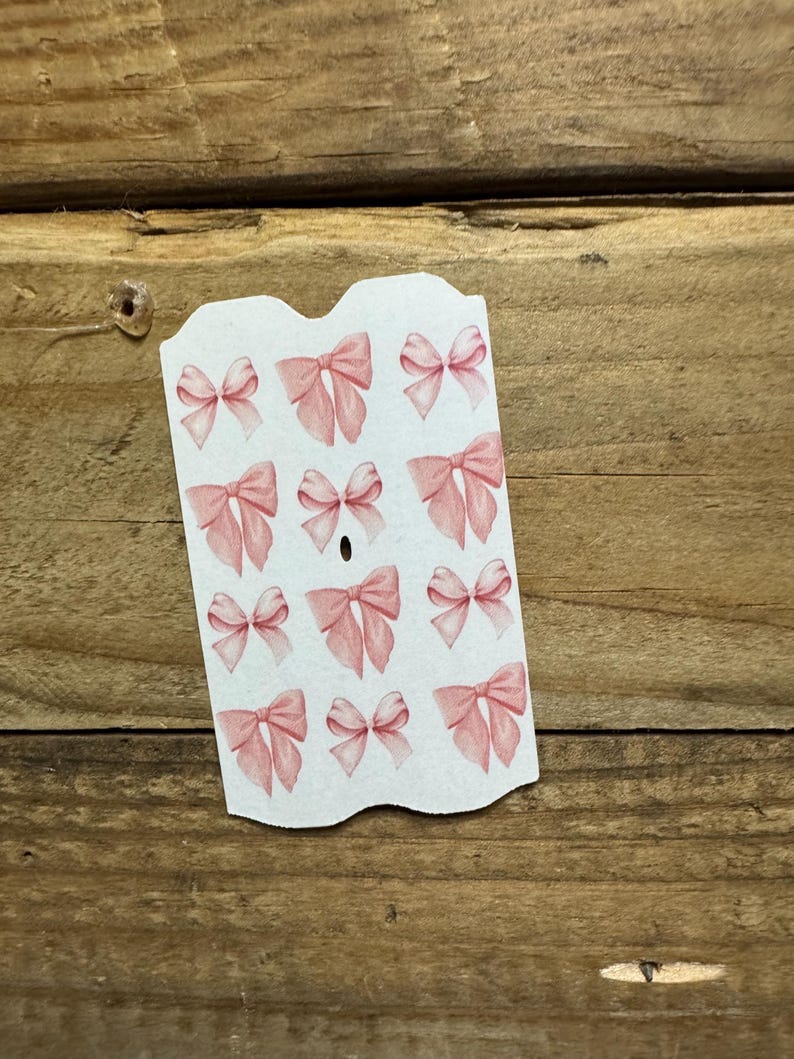 May include: A sheet of stickers featuring watercolor-style pink bows. The bows are in various sizes and arrangements, with a soft, delicate appearance. The stickers are on a white background with a scalloped edge.