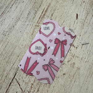 May include: A pink gift tag with a repeating pattern of hearts, bows, and the word "LOVE". The tag has a scalloped edge and is decorated with small hearts and stars. The background is a light pink color.