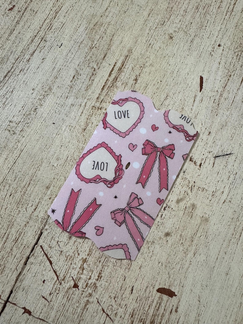 May include: A pink gift tag with a repeating pattern of hearts, bows, and the word "LOVE". The tag has a scalloped edge and is decorated with small hearts and stars. The background is a light pink color.