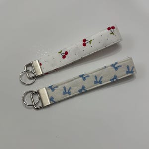 Puede incluir: Dos llaveros de tela. Uno es blanco con cerezas rojas y tallos verdes, el otro es blanco con lazos azules. Cada uno tiene un cierre y un llavero de metal plateado.