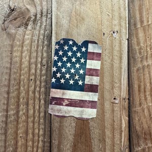 May include: A distressed-look American flag design on a wooden surface. The flag features white stars on a blue background and red and white stripes. The flag is cut in a unique shape.