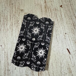 May include: Black and white fabric with a celestial design featuring suns, moons, leaves, and diamond shapes. The fabric has a dark background and a repeating pattern. The edges of the fabric are curved.