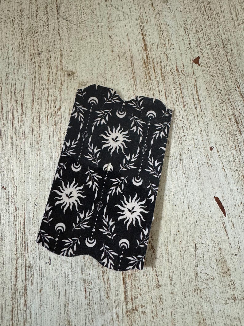May include: Black and white fabric with a celestial design featuring suns, moons, leaves, and diamond shapes. The fabric has a dark background and a repeating pattern. The edges of the fabric are curved.