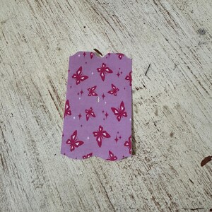 May include: A small, purple tag with a scalloped edge. The tag features a pattern of red butterflies and small diamond shapes. The tag has a small hole at the top.