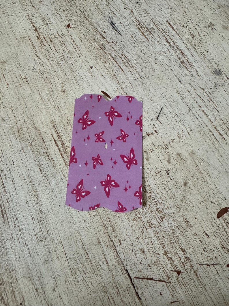 May include: A small, purple tag with a scalloped edge. The tag features a pattern of red butterflies and small diamond shapes. The tag has a small hole at the top.