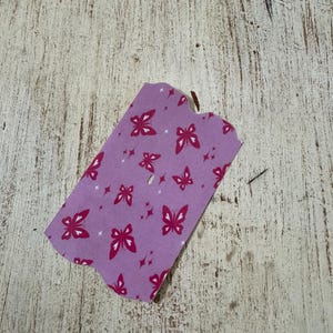 May include: A small, lavender-coloured tag with a butterfly pattern. The butterflies are a deep pink colour and are scattered across the tag. The tag is on a weathered, light-coloured wooden surface.