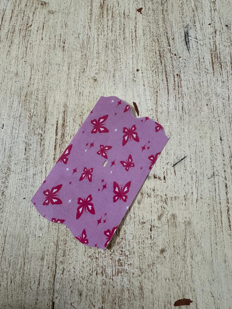 May include: A small, lavender-coloured tag with a butterfly pattern. The butterflies are a deep pink colour and are scattered across the tag. The tag is on a weathered, light-coloured wooden surface.