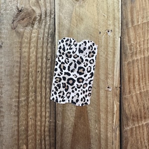 K&ouml;nnte beinhalten: Ein kleiner, rechteckiger Stoffartikel mit Leopardenmuster. Der Druck zeigt schwarze und braune Flecken auf wei&szlig;em Hintergrund. Der Artikel hat eine wellenf&ouml;rmige Oberkante und wird vor einem Holzgrund gezeigt.