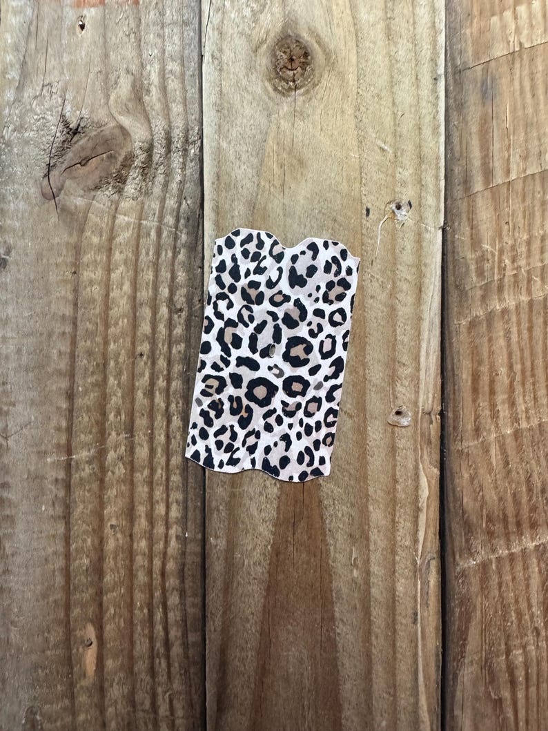 K&ouml;nnte beinhalten: Ein kleiner, rechteckiger Stoffartikel mit Leopardenmuster. Der Druck zeigt schwarze und braune Flecken auf wei&szlig;em Hintergrund. Der Artikel hat eine wellenf&ouml;rmige Oberkante und wird vor einem Holzgrund gezeigt.