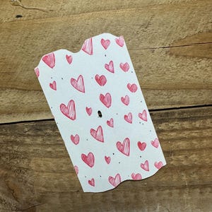Puede incluir: Una etiqueta blanca con borde festoneado est&aacute; cubierta de garabatos de corazones rojos. Los corazones var&iacute;an en tama&ntilde;o y est&aacute;n dispersos por la etiqueta. La etiqueta tiene un peque&ntilde;o agujero cerca de la parte superior. El fondo es una superficie de madera.