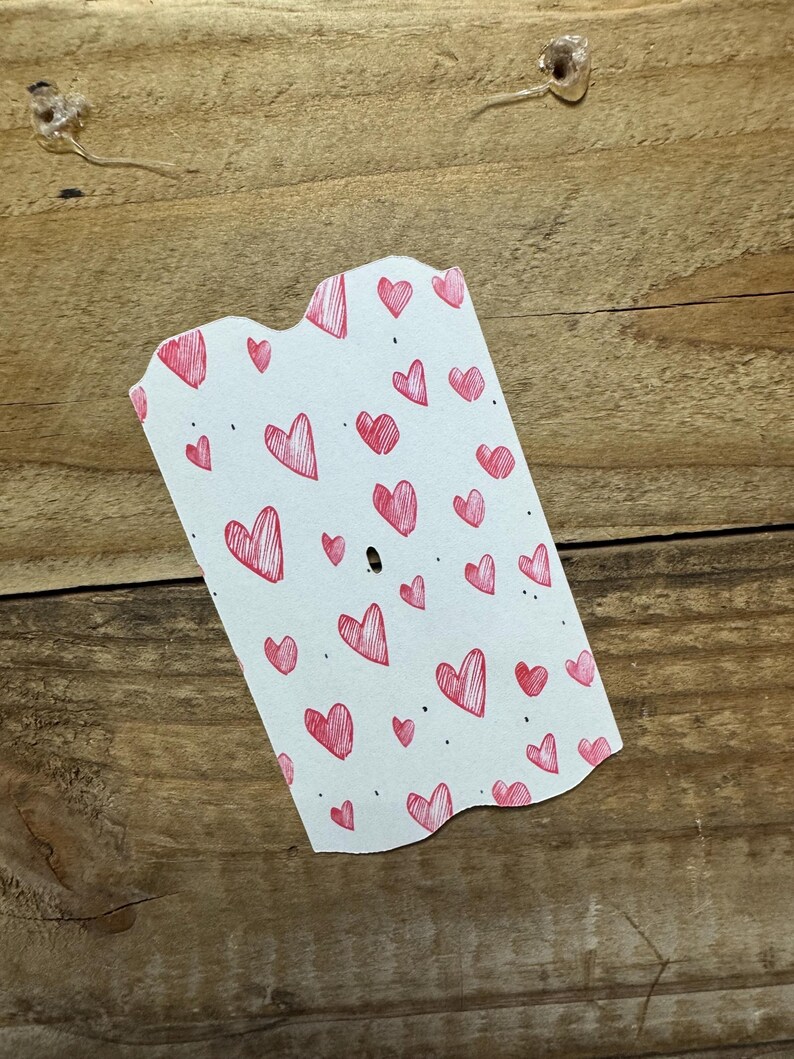Puede incluir: Una etiqueta blanca con borde festoneado est&aacute; cubierta de garabatos de corazones rojos. Los corazones var&iacute;an en tama&ntilde;o y est&aacute;n dispersos por la etiqueta. La etiqueta tiene un peque&ntilde;o agujero cerca de la parte superior. El fondo es una superficie de madera.