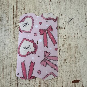 May include: A pink card with a repeating pattern of hearts with the word "LOVE", bows, and small hearts. The card has a scalloped edge and is set against a distressed, light-colored wooden surface.