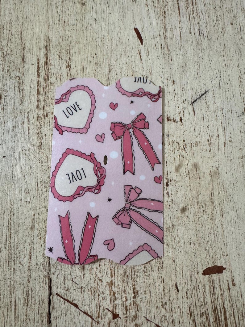 May include: A pink card with a repeating pattern of hearts with the word "LOVE", bows, and small hearts. The card has a scalloped edge and is set against a distressed, light-colored wooden surface.