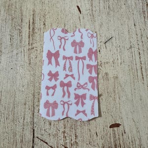 Op de afbeelding: Een wit label met een geschulpte rand met een patroon van roze strikken in verschillende stijlen. De strikken zijn gedrukt op een witte achtergrond. Het label ligt op een lichtbruin, getextureerd oppervlak.