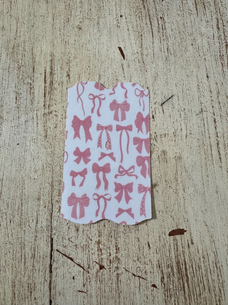 Op de afbeelding: Een wit label met een geschulpte rand met een patroon van roze strikken in verschillende stijlen. De strikken zijn gedrukt op een witte achtergrond. Het label ligt op een lichtbruin, getextureerd oppervlak.