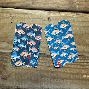 May include: Two decorative tags with a fish pattern. One tag has a dark blue background with orange and blue fish. The other tag has a light blue background with orange and white fish. The number "1" is written on the first tag and the number "2" is written on the second tag.