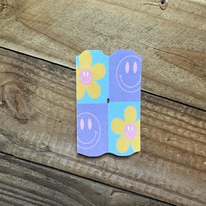May include: A small, folded paper card with a repeating pattern of smiley faces and flowers. The card features a light blue, yellow, and purple color scheme. The background is a wooden surface.