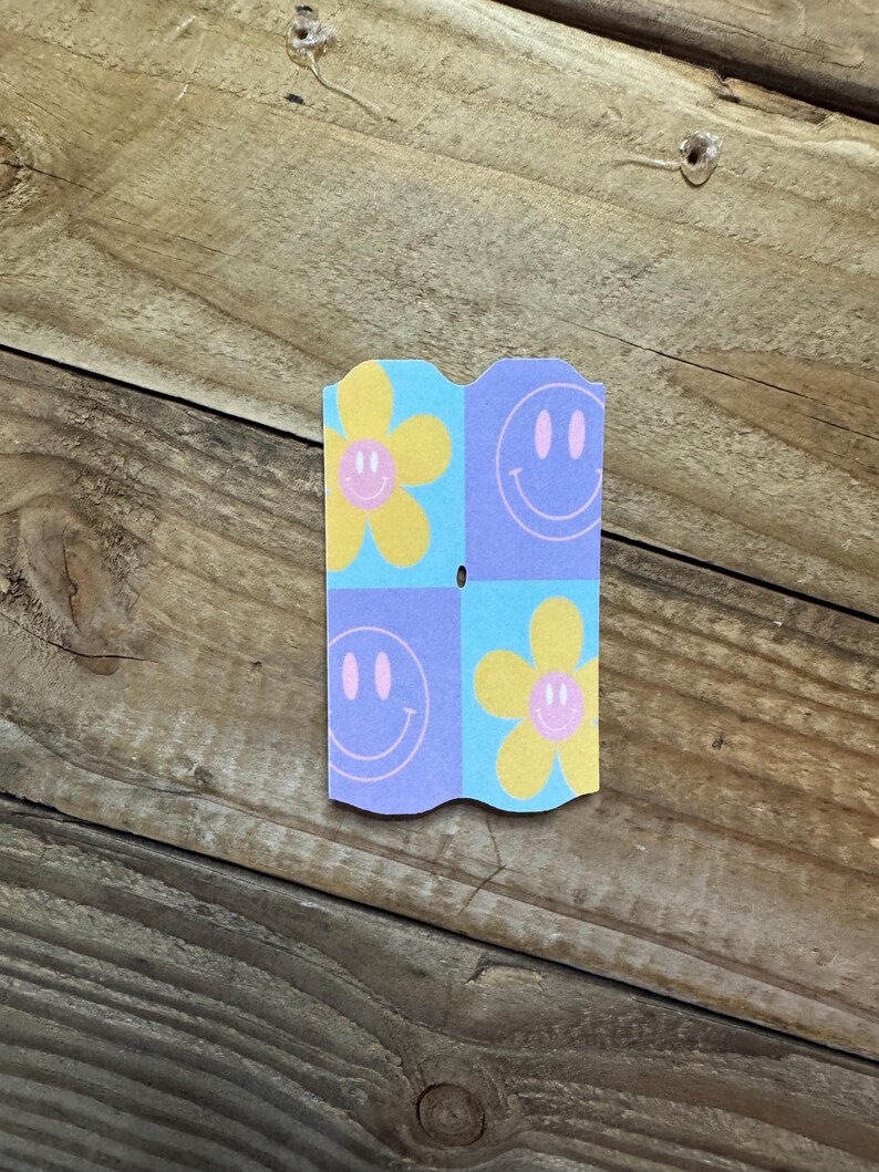 May include: A small, folded paper card with a repeating pattern of smiley faces and flowers. The card features a light blue, yellow, and purple color scheme. The background is a wooden surface.
