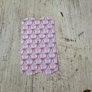 Puede incluir: Una etiqueta rosa y blanca con estampado de cuadros vichy y borde superior festoneado. La etiqueta est&aacute; decorada con un patr&oacute;n repetido de lazos rosas. La etiqueta est&aacute; sobre una superficie de madera clara y desgastada.