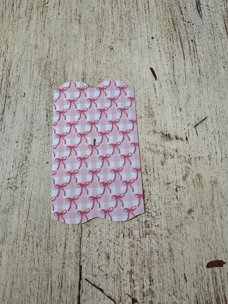 Puede incluir: Una etiqueta rosa y blanca con estampado de cuadros vichy y borde superior festoneado. La etiqueta est&aacute; decorada con un patr&oacute;n repetido de lazos rosas. La etiqueta est&aacute; sobre una superficie de madera clara y desgastada.