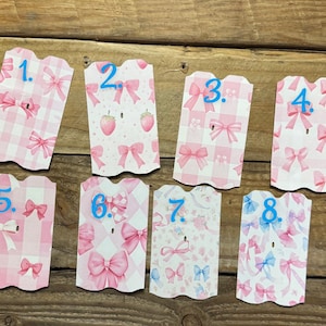 May include: Eight decorative gift tags with scalloped edges. The tags feature various pink and blue bow and strawberry patterns. Each tag is numbered in blue. The tags are arranged on a wooden surface.